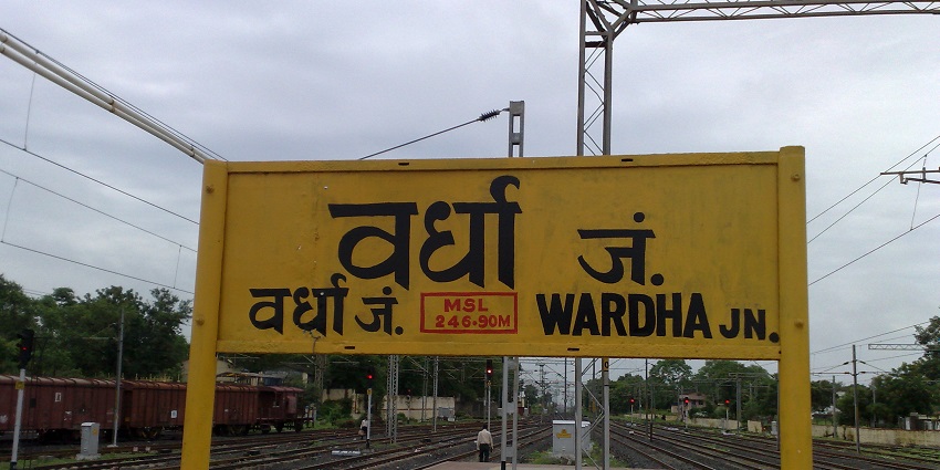 A railway platform at Wardha Junction with a train on the tracks and a station sign visible.