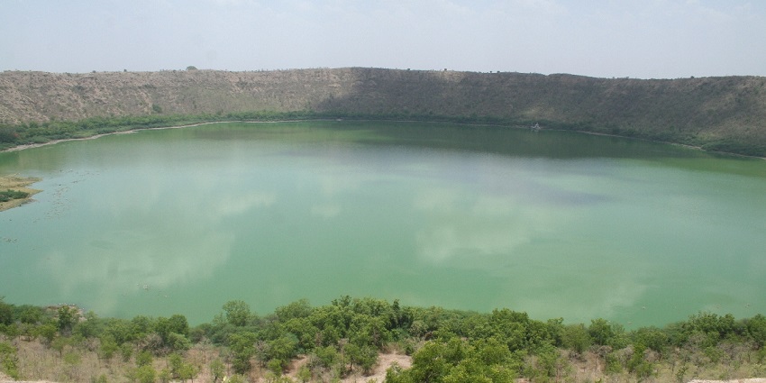 A large, circular lake surrounded by hills, with green water and lush vegetation.