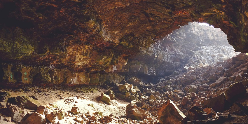A narrow brown tunnel leads to a bright exit, with water flowing underneath and stone walls
