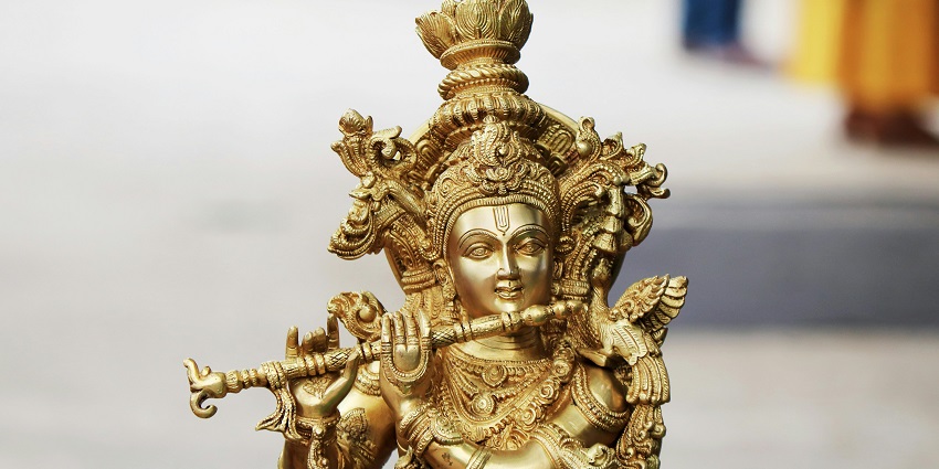 A picture of a golden idol of Lord Krishna with a scenic background filled with trees and greenery