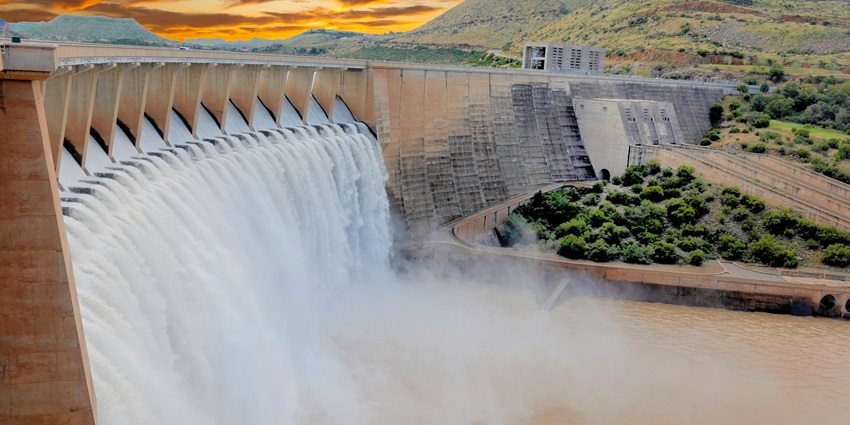 A scenic view of a Dam during daytime with calm water and a forested hill in the background