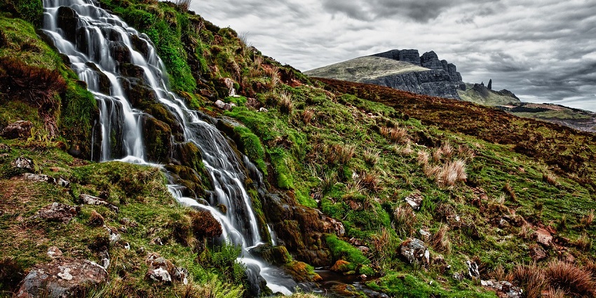 Mhaismal Waterfall is a small waterfall trickling down a rocky mountain slope surrounded by green trees and vegetation.