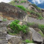 Ancient stone ruins of the Morgiri Fort sit on a mountain with green vegetation and a cloudy sky in the background