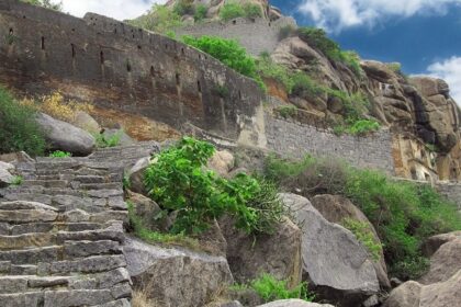 Ancient stone ruins of the Morgiri Fort sit on a mountain with green vegetation and a cloudy sky in the background