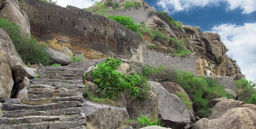 Ancient stone ruins of the Morgiri Fort sit on a mountain with green vegetation and a cloudy sky in the background