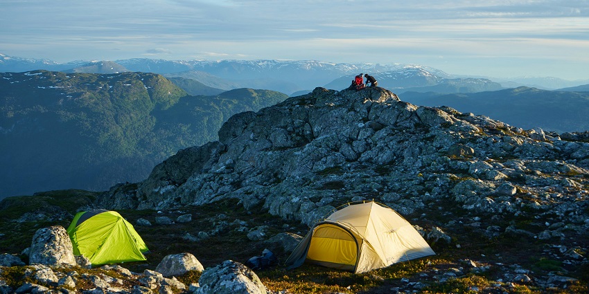 A distant view of two people sitting on a mountain peak near which tents are placed