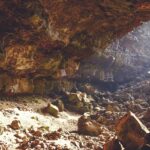 Sunlight peeks through the upper hollow of a cave, with rocks of various sizes inside.