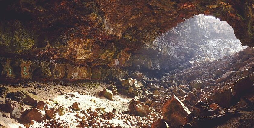 Sunlight peeks through the upper hollow of a cave, with rocks of various sizes inside.