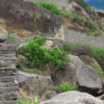 Ancient stone ruins sit on a mountain, surrounded by lush greenery under a cloudy sky.