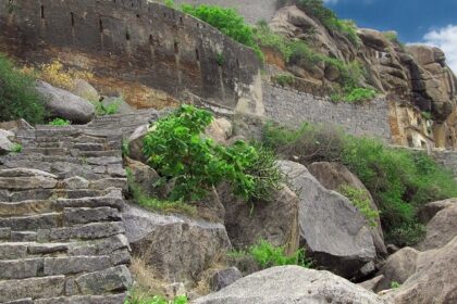 Ancient stone ruins sit on a mountain, surrounded by lush greenery under a cloudy sky.