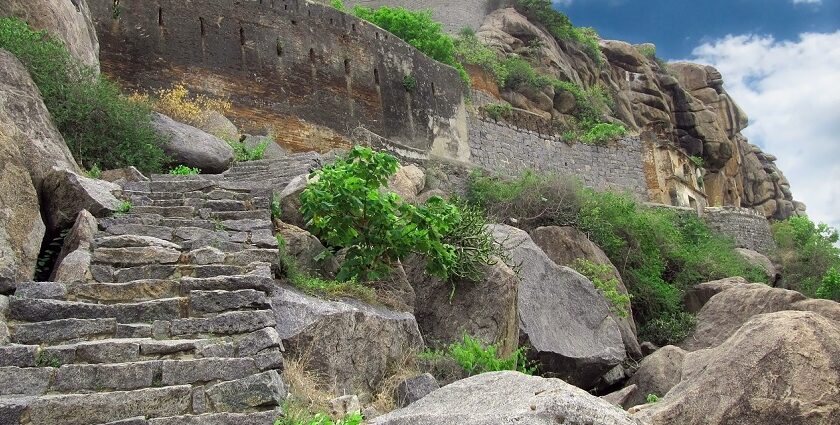 Ancient stone ruins sit on a mountain, surrounded by lush greenery under a cloudy sky.