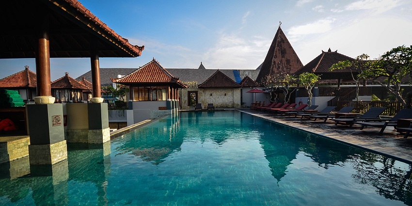 A modern resort building with a large swimming pool, surrounded by palm trees under a clear blue sky.