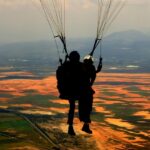 Silhouette of tandem paragliders flying above green and glowing horizon during sunset