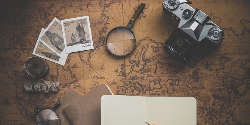 A camera, notebook, pencil, spectacles and more displayed over a map on a table for travel planning.