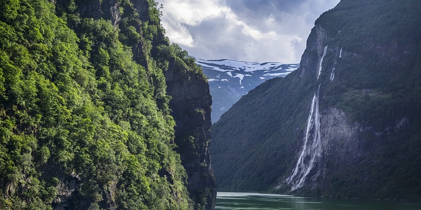 A waterfall flows down rocky cliffs into a river, surrounded by green trees and misty mountain peaks.