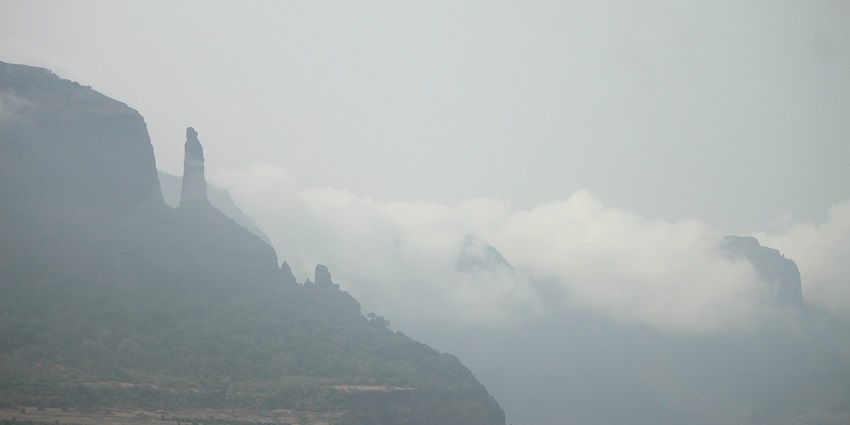 A steep rocky cliff with scattered greenery, leading to the remains of Jivdhan Fort under a cloudy sky.