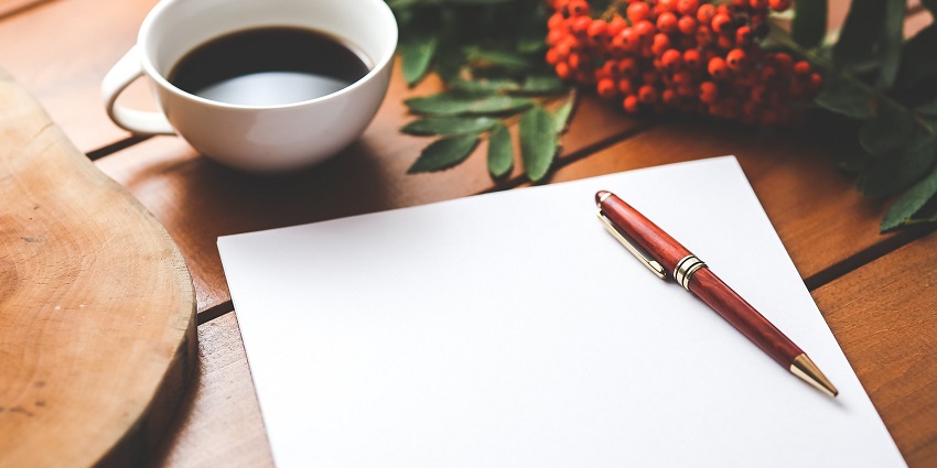 An aerial shot of a white paper on a brown table with a cup of coffee on the side.