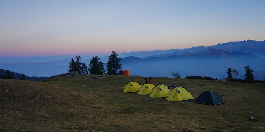 Tents set on the grassy terrain campsite of Bhoj Gadi, surrounded by clouds and mountains
