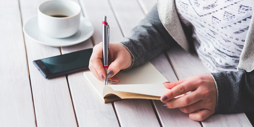 A view of a woman with a pen in her hands writing on a white paper with a cup of coffee.