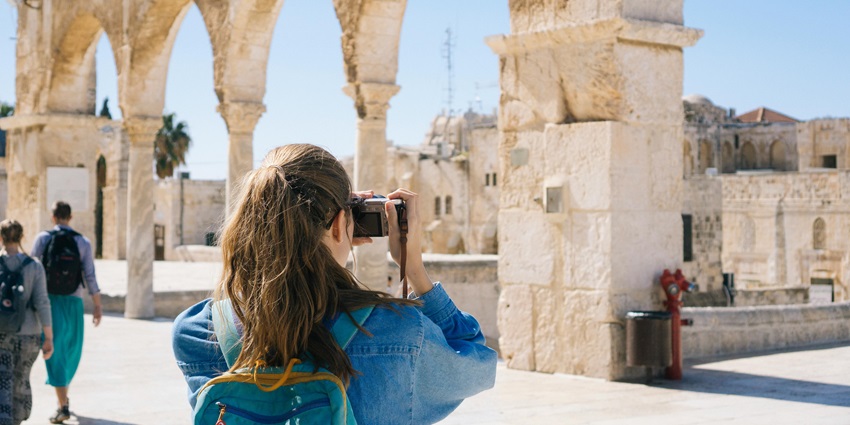 Woman photographing ancient ruins surrounded by stone structures under a bright, clear sky