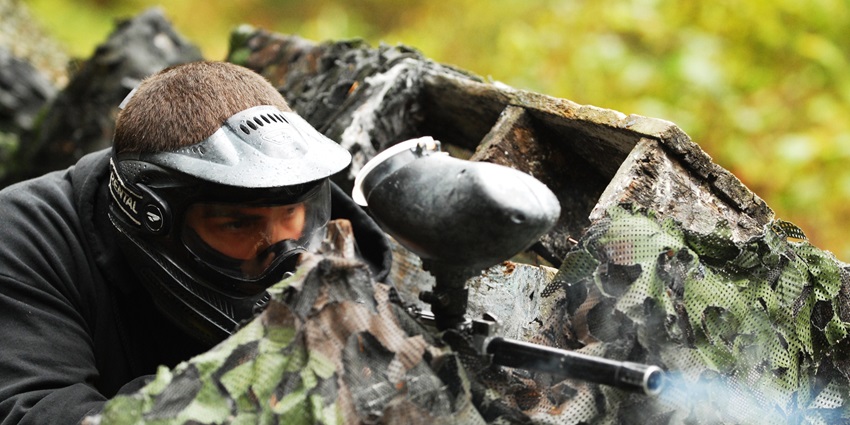 An image of a person with a paintball gun, hiding behind a cover and firing.
