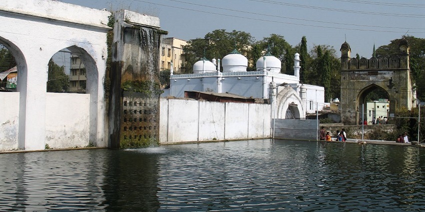 A stone fountain at Panchakki, Aurangabad, with water flowing into a large, round pool surrounded by trees.