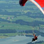 A paraglider waving while soaring high above rugged mountains and valleys with lakes.