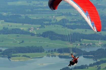 A paraglider waving while soaring high above rugged mountains and valleys with lakes.