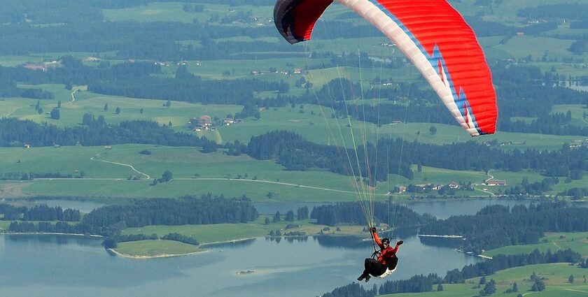 A paraglider waving while soaring high above rugged mountains and valleys with lakes.