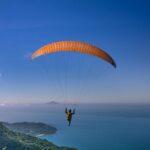 Person solo paragliding mid-air surrounded by green mountains and a vast sea below.