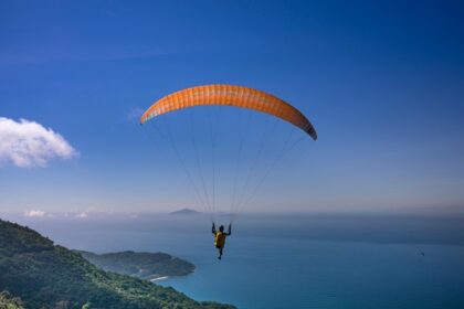 Person solo paragliding mid-air surrounded by green mountains and a vast sea below.