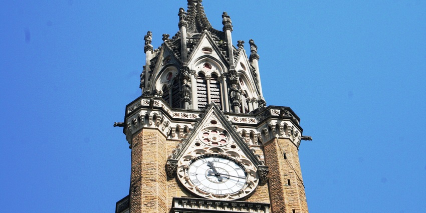 A picture of the top of a clock tower in Mumbai with a massive clock in the centre