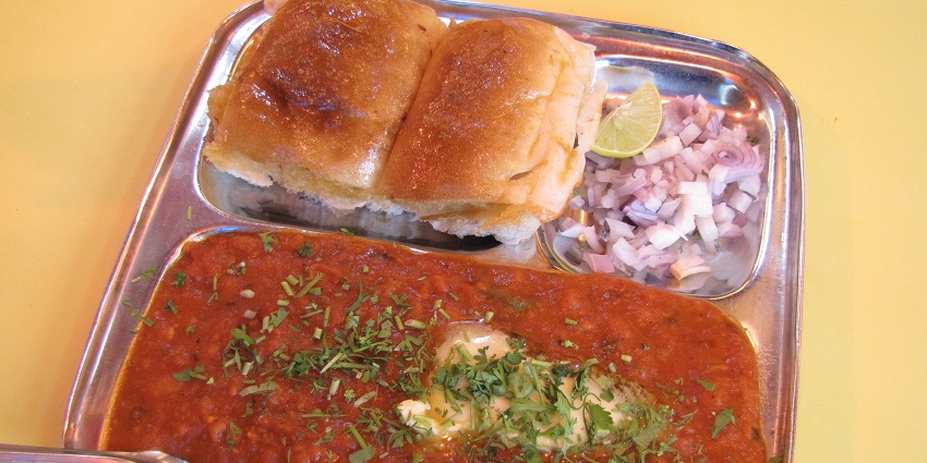 Buttery red bhaji served with buttery pav, chopped onions and a lime wedge on a steel plate.