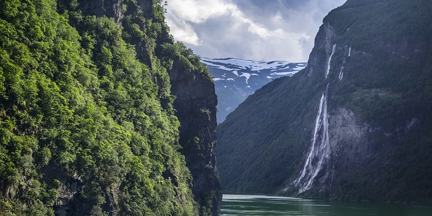 Sakhar Kheda Waterfall flows down rocky cliffs into a river, surrounded by green trees and misty mountain peaks.