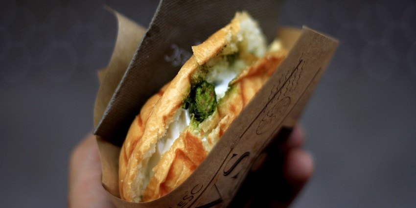 A person holding a grilled cheese sandwich stuffed with greens, wrapped in brown paper.