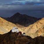 A picture of a monastery perched on top of a hill in Leh with multiple mountains visible in the background