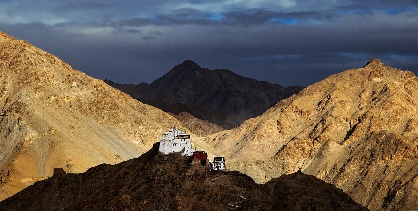 A picture of a monastery perched on top of a hill in Leh with multiple mountains visible in the background