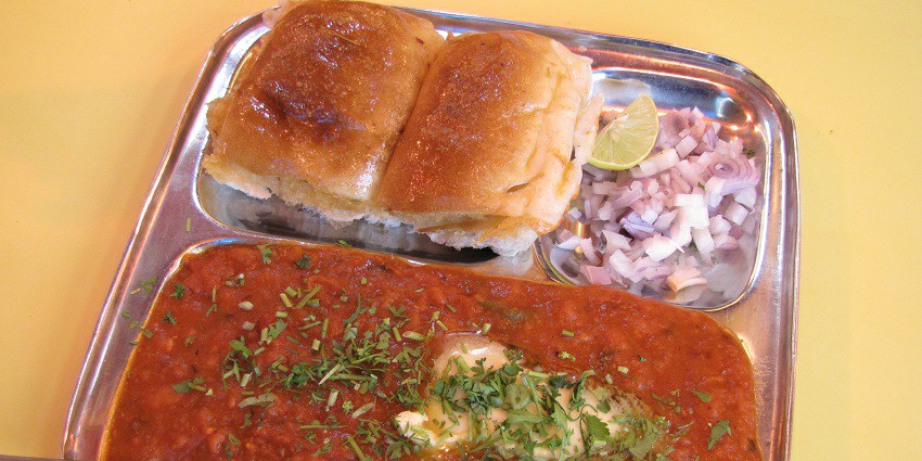 Bhaji served with buttered pavs, chopped onions and half lemon on a stainless steel plate
