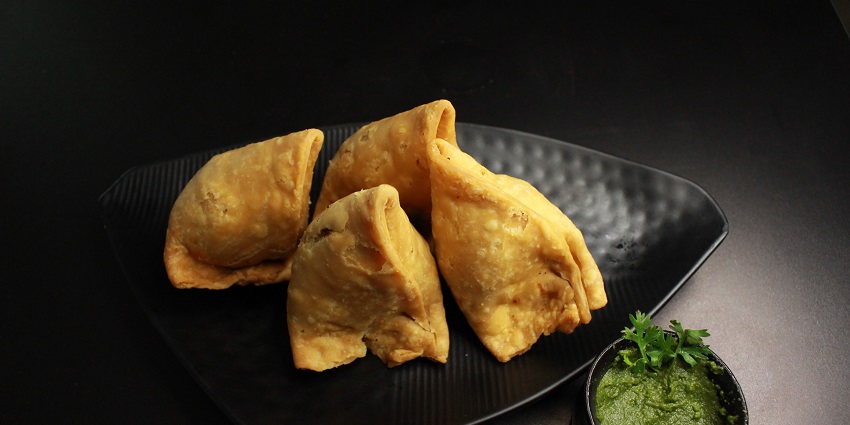 Four pieces of samosas served on a black plate accompanied by green coriander chutney.