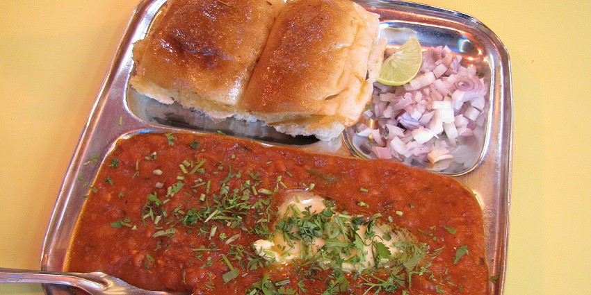 Buttery red bhaji served with buttery pav, chopped onions and lime wedge on a steel plate.