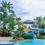 People enjoying a large swimming pool with slides and water features in a park setting.