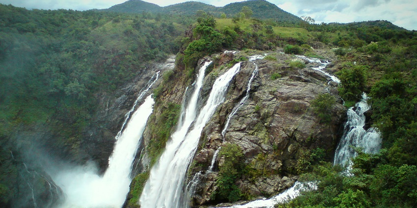 Shivanasamudra Falls cascades over cliffs, surrounded by vibrant foliage and rocks.