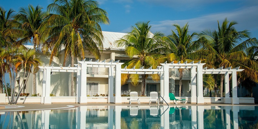 A luxury hotel with palm trees, a pool, and lounge chairs under a bright blue sky and sunny weather.