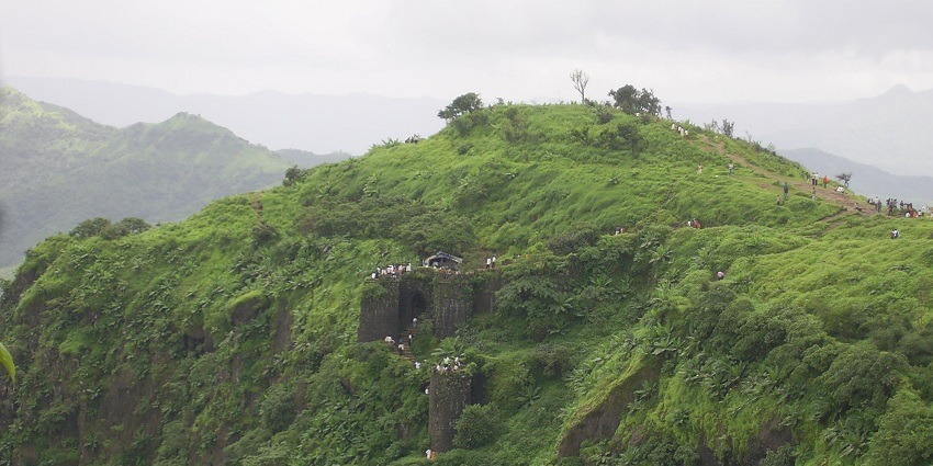 A scenic view of Sinhagad Fort on a hilltop, surrounded by greenery and partially cloudy skies.