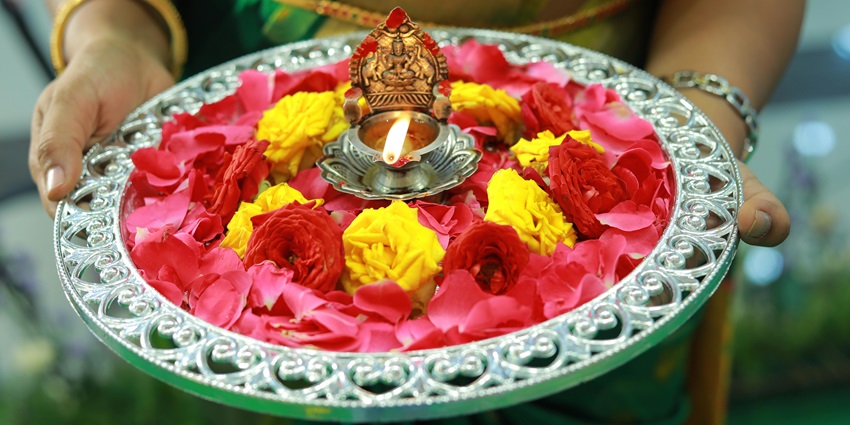 Image of a devotee holding a pooja thali decorated with colourful flowers and a diya in the middle