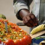 A man decoratively cutting a raw mango among colourful display of veggies in a food stall