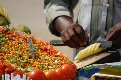 A man decoratively cutting a raw mango among colourful display of veggies in a food stall