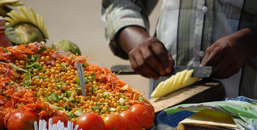 A man decoratively cutting a raw mango among colourful display of veggies in a food stall