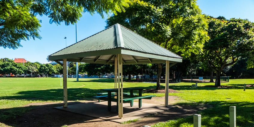 A shaded picnic shelter with benches in a sunny park, surrounded by lush green grass.