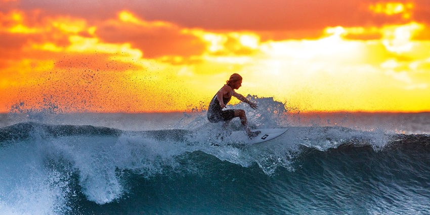 An image showing surfers riding a wave during sunset.